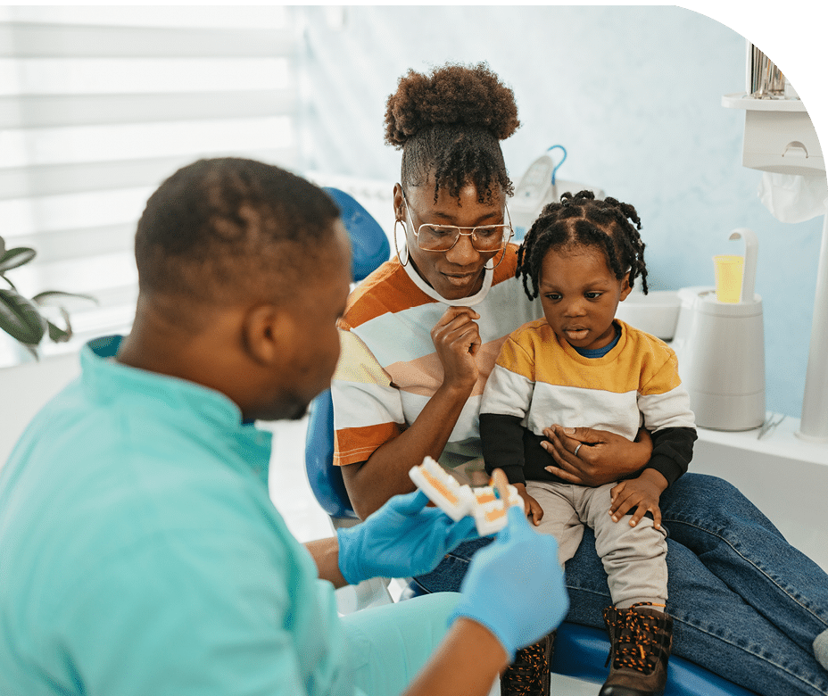 African american other and her son visiting dentist office, she is sitting in the dentist chair while her son is in the lap Doctor has plastic teeth model and is explaining how to brush teeth