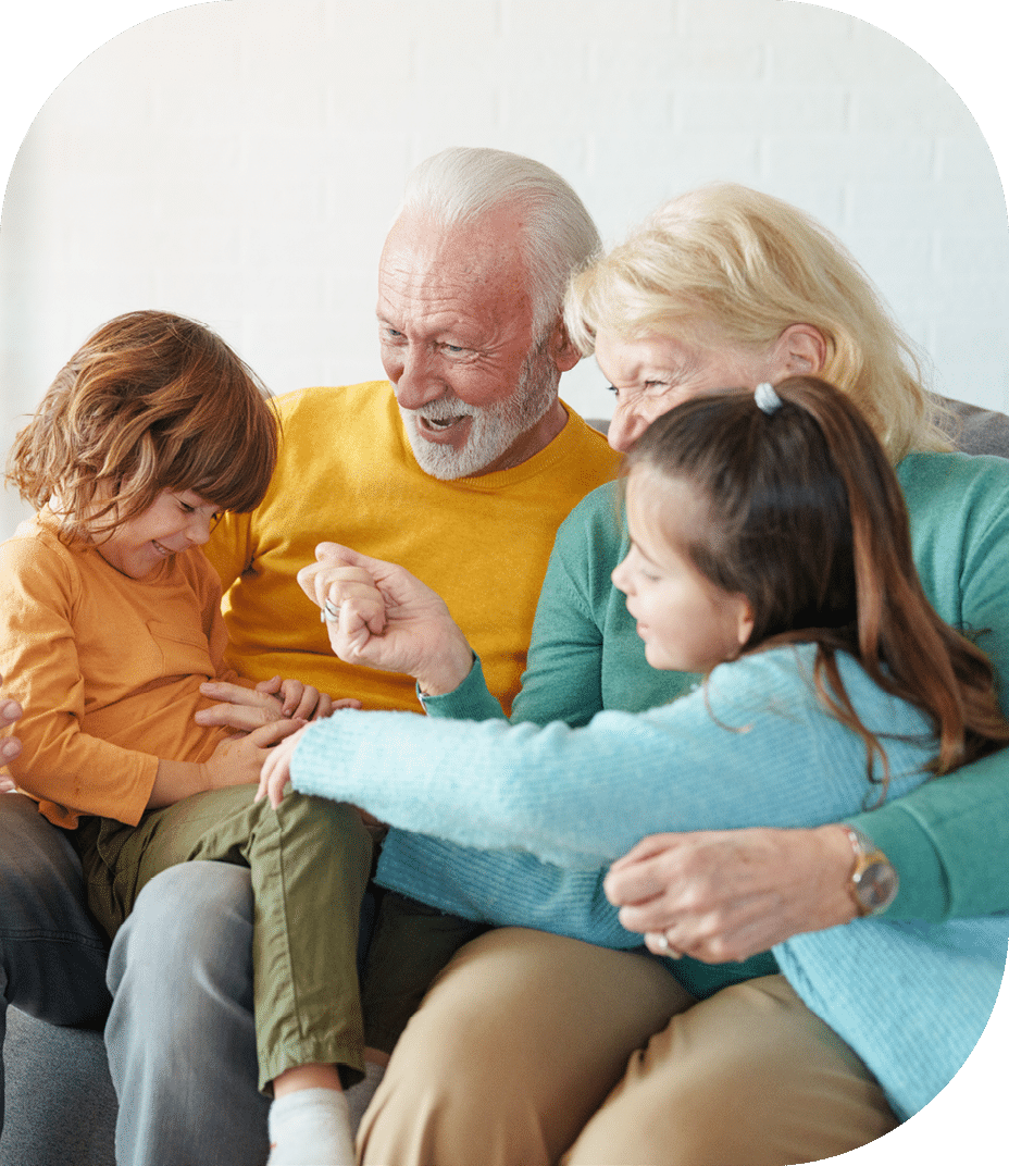 Candid portrait of Caucasian senior couple in casual attire sharing joyful interaction with two young grandchildren on gray sofa in naturally lit modern living space.